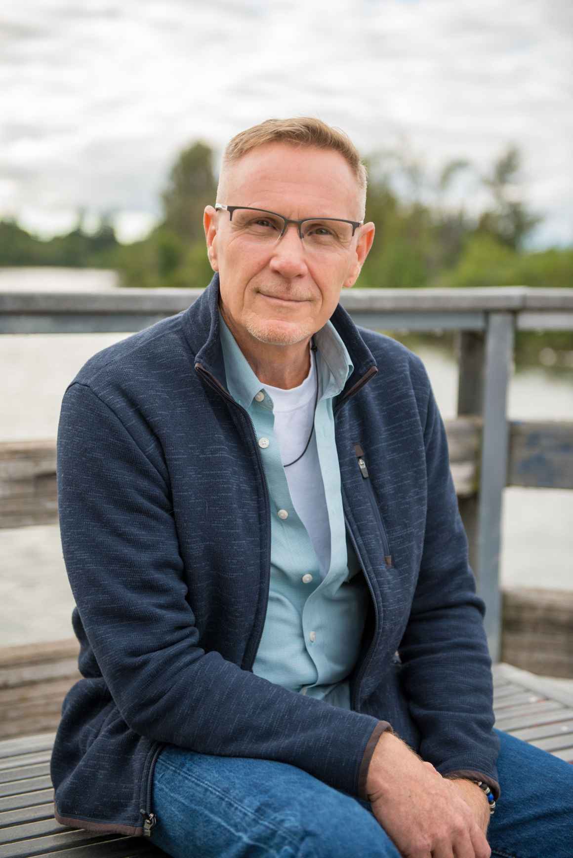 A portrait of our client, John Cavanaugh, a middle aged white man wearing glasses, a blue button up, and a gray cardigan, sitting on a bench with his hands clasped, softly smiling