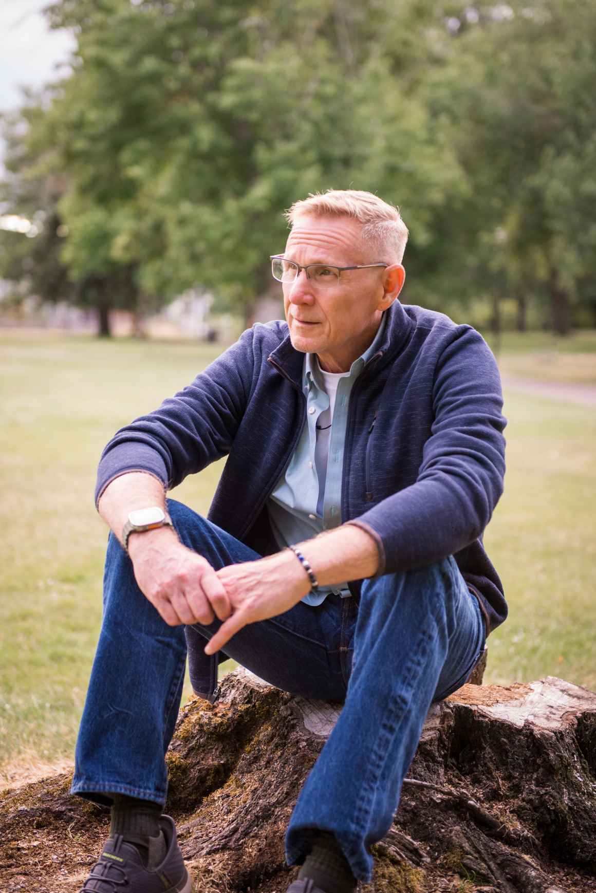 ACLU client, John Cavanaugh, a middle aged white man, sits on a tree stump looking away from camera, wearing glasses, a gray cardigan and jeans.