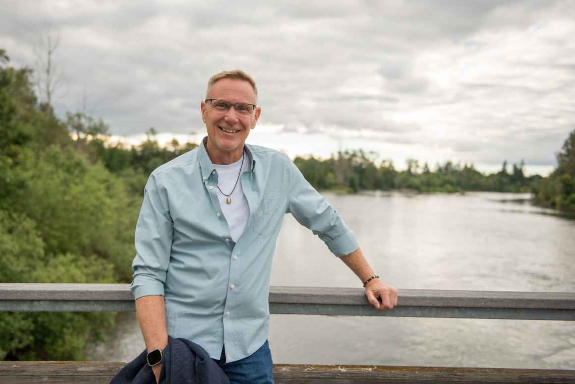 ACLU client, John Cavanaugh, a middle aged white man, smiles at the camera while standing in front of a river on a bridge. He is wearing glasses and a light blue button up shirt.