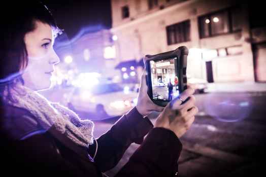 Carrie Medina holds a tablet to film the police on the street. Police lights blur into the foreground.