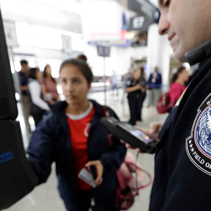 A passenger using a facial recognition kiosk in the background with a U.S. Customs and Border Protection officer watching in the foreground.